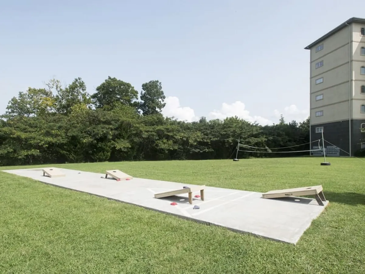 An active, open recreational area with bean bag toss boards, a volleyball net, a grassy field, and gathering spaces surrounded by trees and a building in the background. The setting is perfect for group games and socializing.
