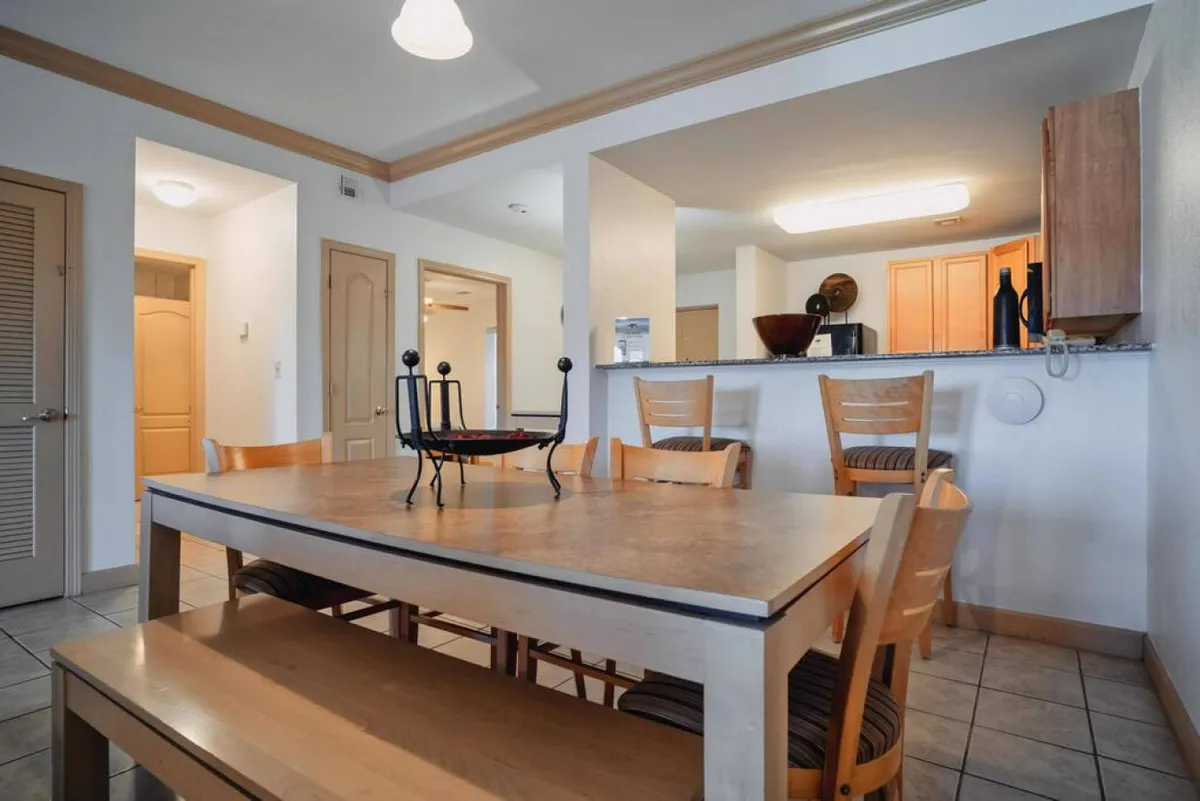 An open-plan dining area seamlessly connected to the kitchen, with a wooden dining table and chairs, decorative bowl, and abundant natural light. The arrangement encourages communal meals, conversation, and celebration.