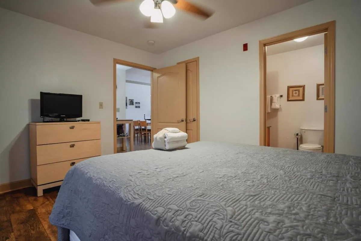 A bedroom with a queen-sized bed, a wooden dresser with a TV, and a doorway connecting to the rest of the home. The space is styled for comfort and privacy, offering a relaxing environment for guests to unwind and enjoy personal downtime.