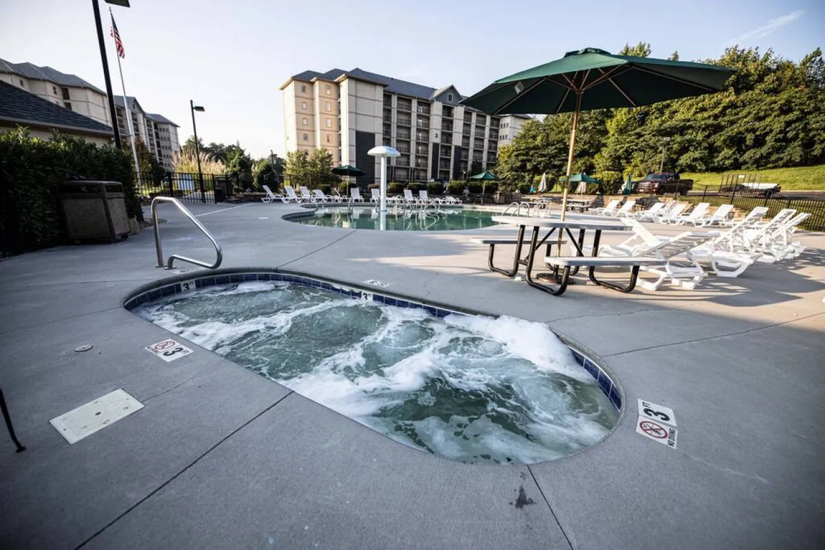 The exterior amenities of the resort are on display, including a sparkling swimming pool, inviting hot tub, several lounge chairs, a shaded picnic table, and a sun umbrella. The area is alive with possibilities for group gatherings, poolside relaxation, and accessible fun for all ages.