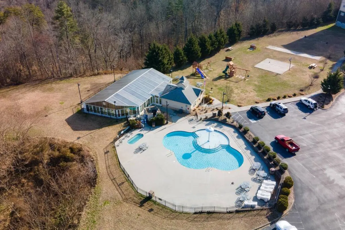 This vibrant exterior shot captures the outdoor swimming pool surrounded by a recreation area, visible playground equipment, and a glass-enclosed building in the background. The scene is bustling with opportunities for swimming, sunbathing, and children’s play, all set within a family-friendly atmosphere.