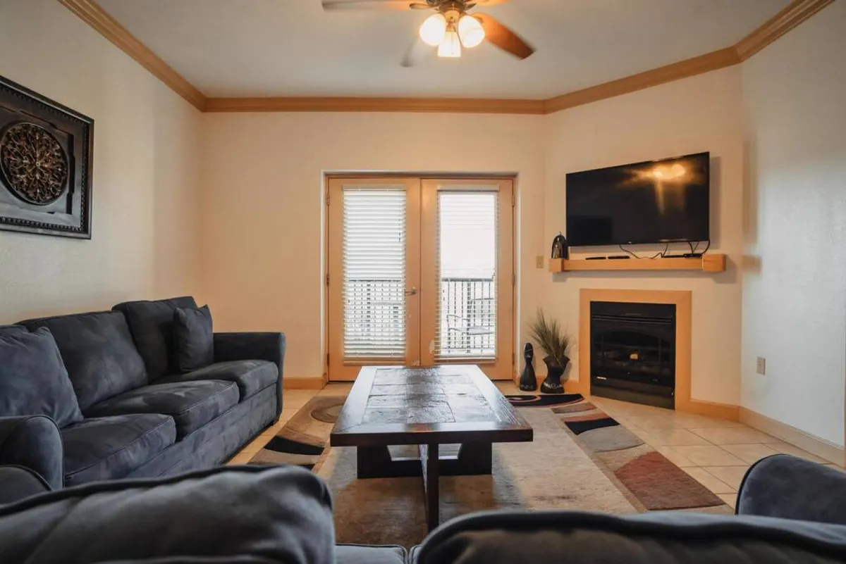 A spacious, bright living room featuring a comfortable sofa set, a sleek coffee table, a fireplace, a large flat screen TV, and balcony doors that invite in natural light. The arrangement encourages relaxation, movie nights, and socializing.