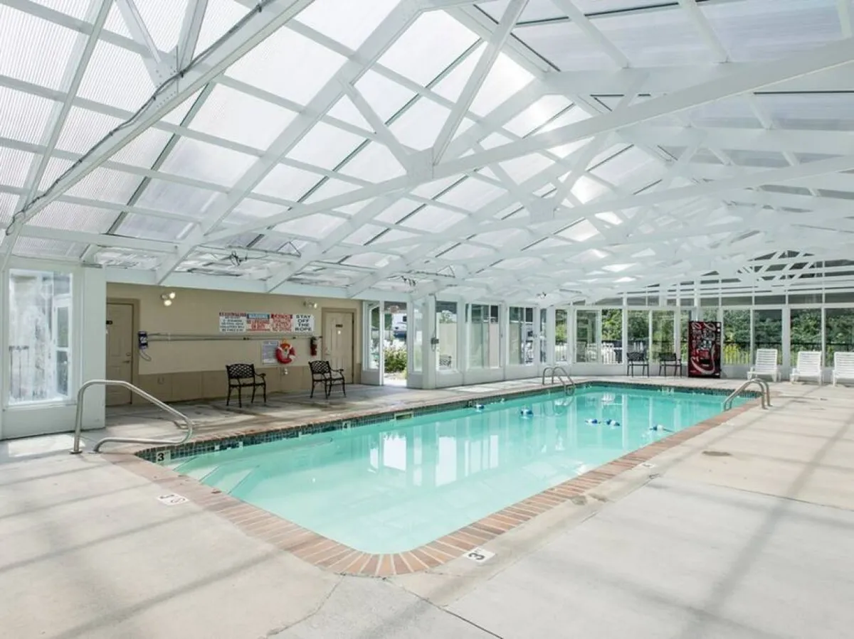 A serene interior space features a spacious indoor pool with windowed walls that let in ample natural light. Poolside seating lines the water, inviting relaxation and year-round enjoyment regardless of weather.